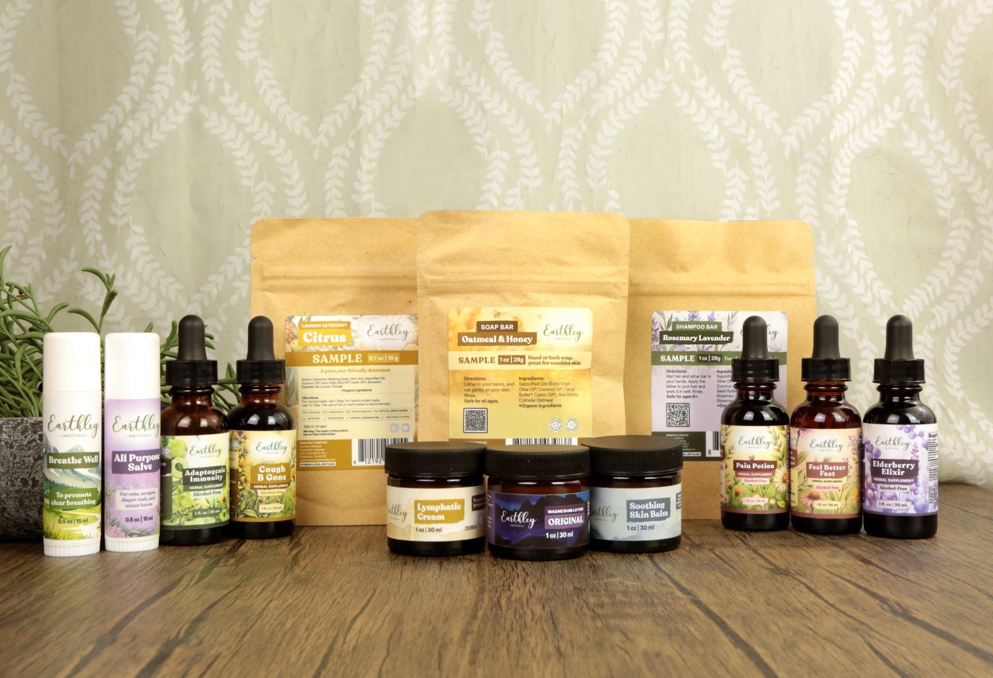 Earthley product collection display showing variety of wellness products including Breathe Well, All Purpose Salve, Adaptogenic Immunity, Cough B Gone tinctures, Citrus sample, Oatmeal and Honey soap bar, Rosemary Lavender shampoo bar, Lymphatic Cream, Magnesium Lotion Original, Soothing Skin Balm, Pain Potion, Feel Better Fast, and Elderberry Elixir arranged on wooden surface