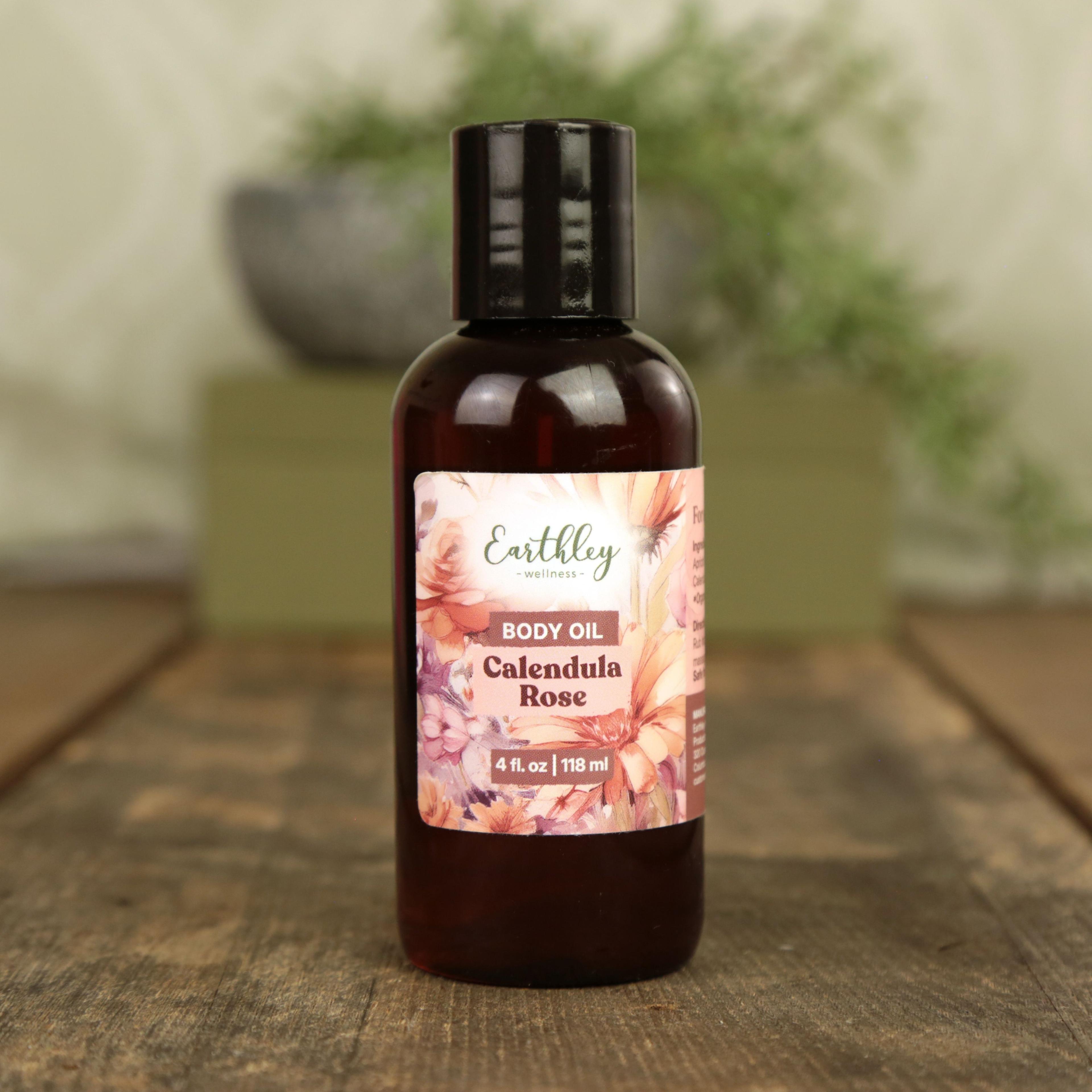 Earthley Calendula Rose Body Oil in 4 oz amber bottle with black cap and pink floral label on wooden surface with greenery in soft focus background