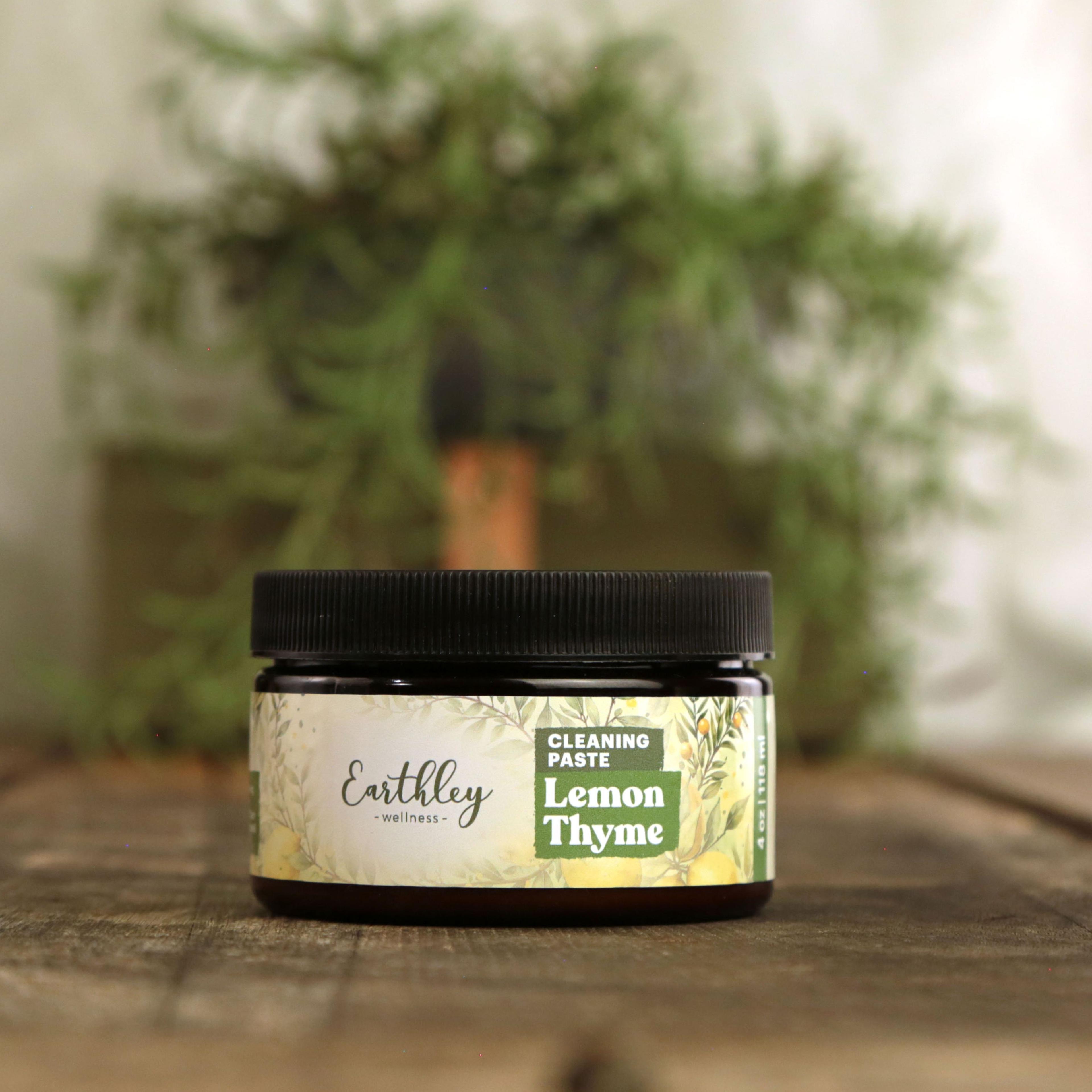 Earthley Cleaning Paste Lemon Thyme 4 oz amber glass jar with black lid featuring botanical label with lemons, thyme, and green herbs, on wooden surface with trailing rosemary plant in background