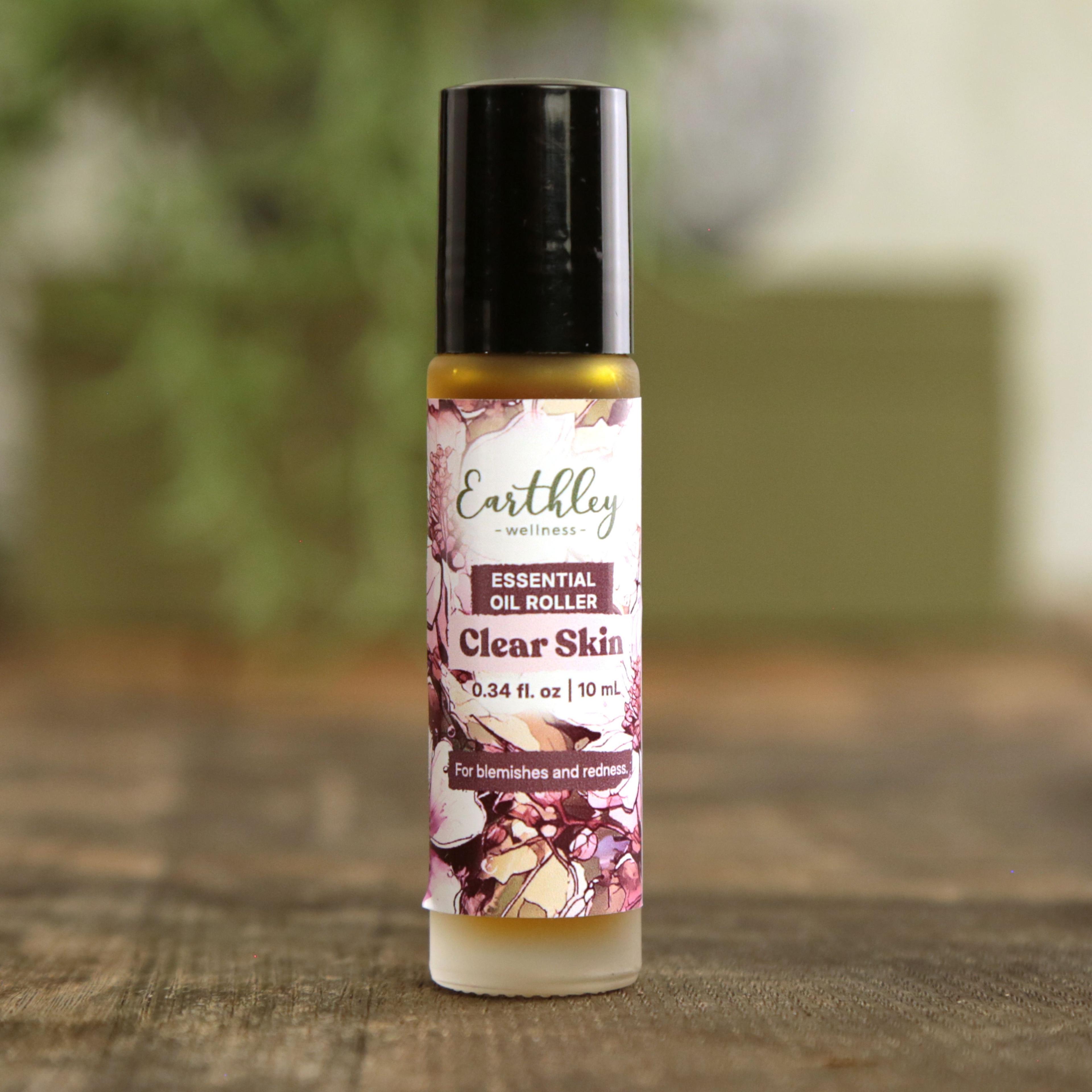 Earthley Clear Skin Essential Oil Roller 0.34 oz glass roll-on bottle with purple and pink abstract floral label, labeled For blemishes and redness, on wooden surface with greenery in background