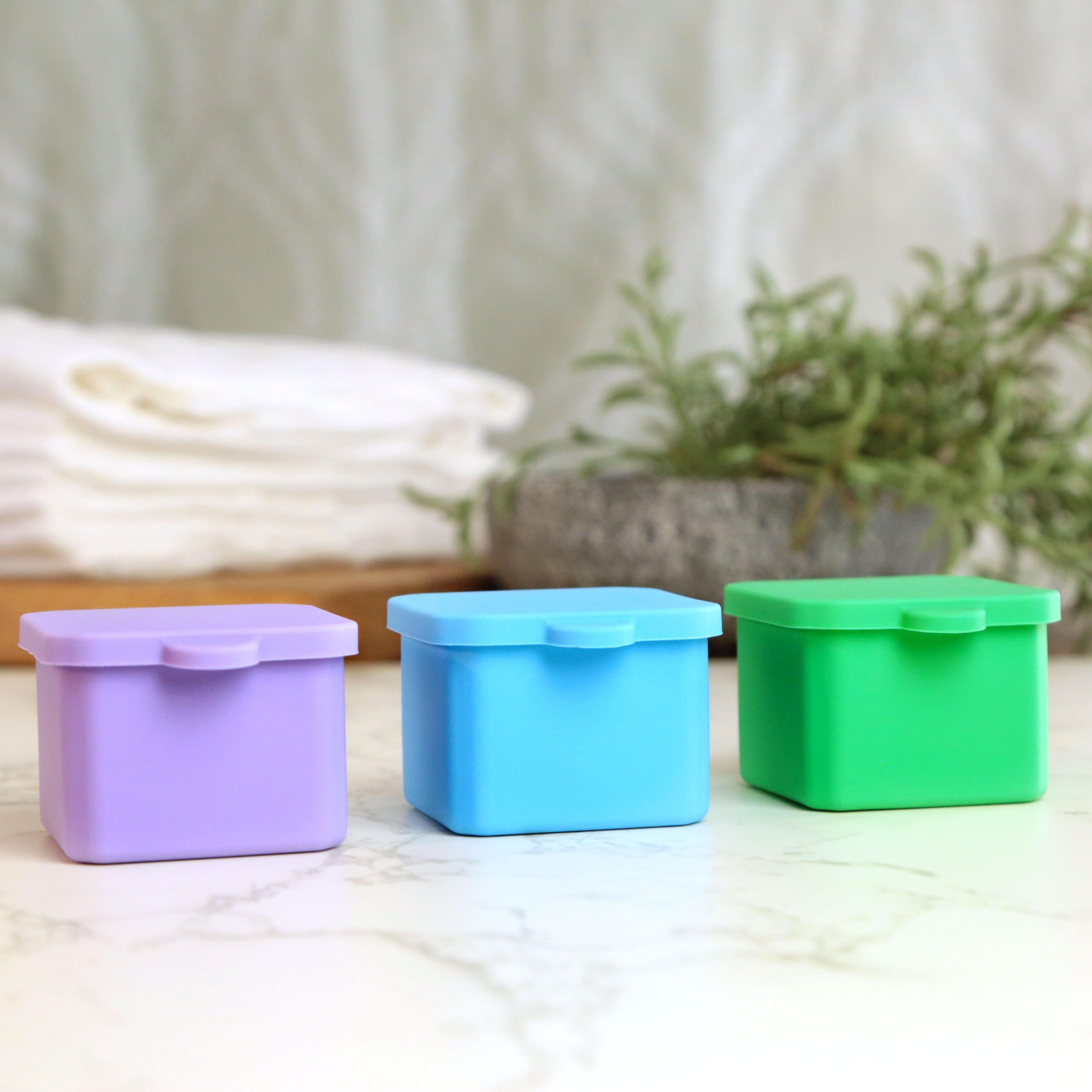 Three silicone storage containers in purple, blue, and green with snap lids on marble surface.
