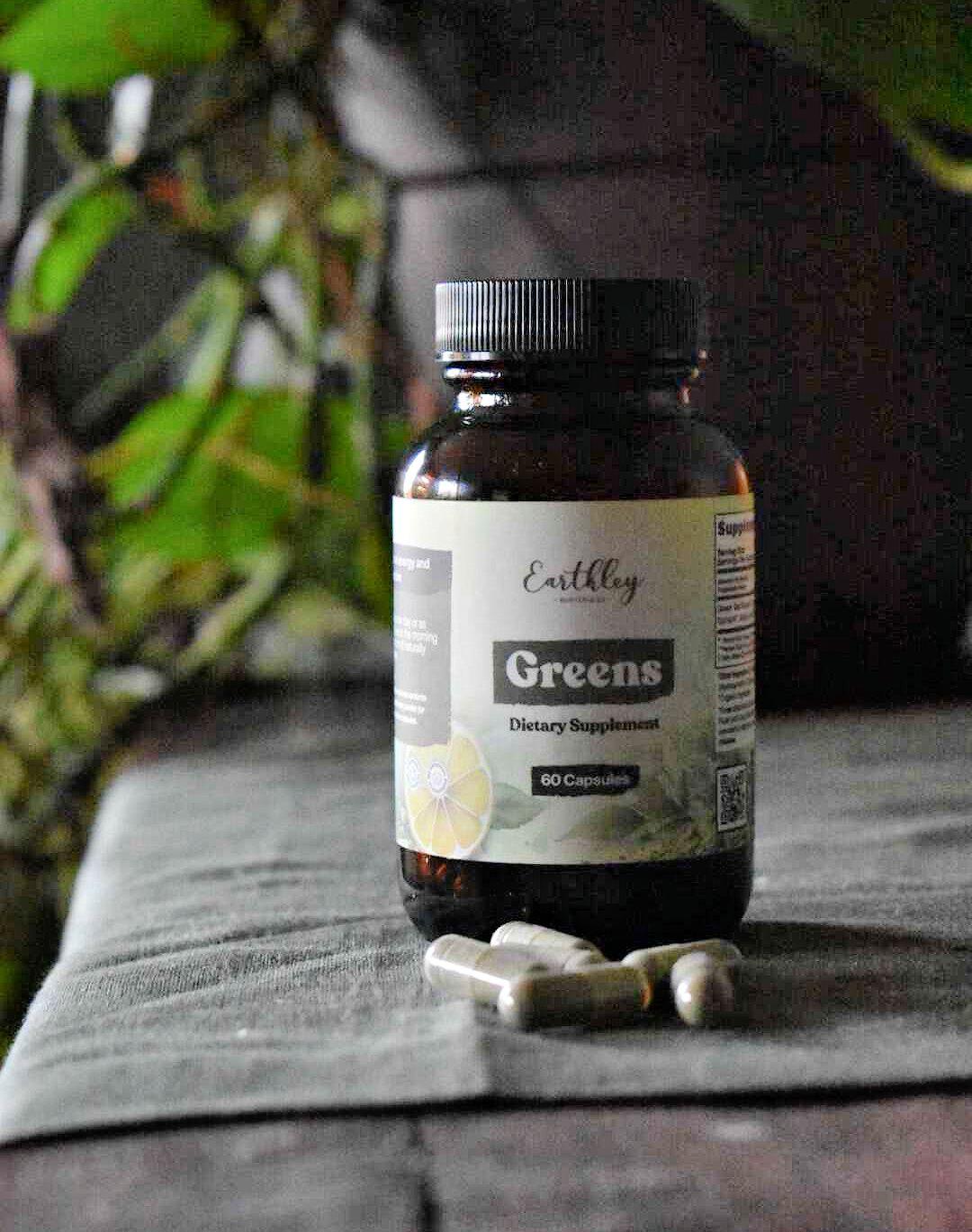 Earthley Greens dietary supplement bottle with 60 capsules, several capsules scattered nearby, green plants in background
