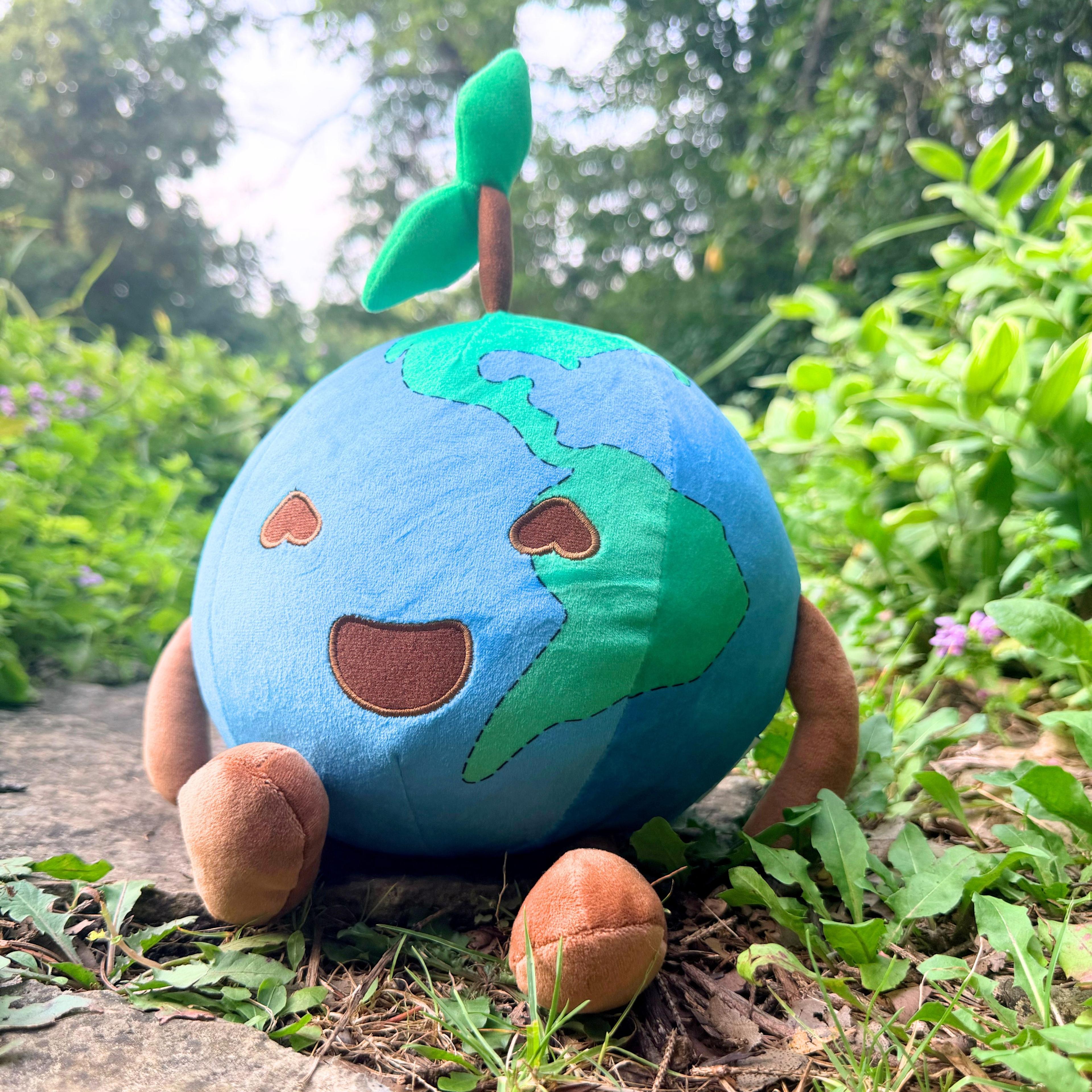 Smiling Earth plush toy with heart eyes and green sprout, sitting outdoors among plants.