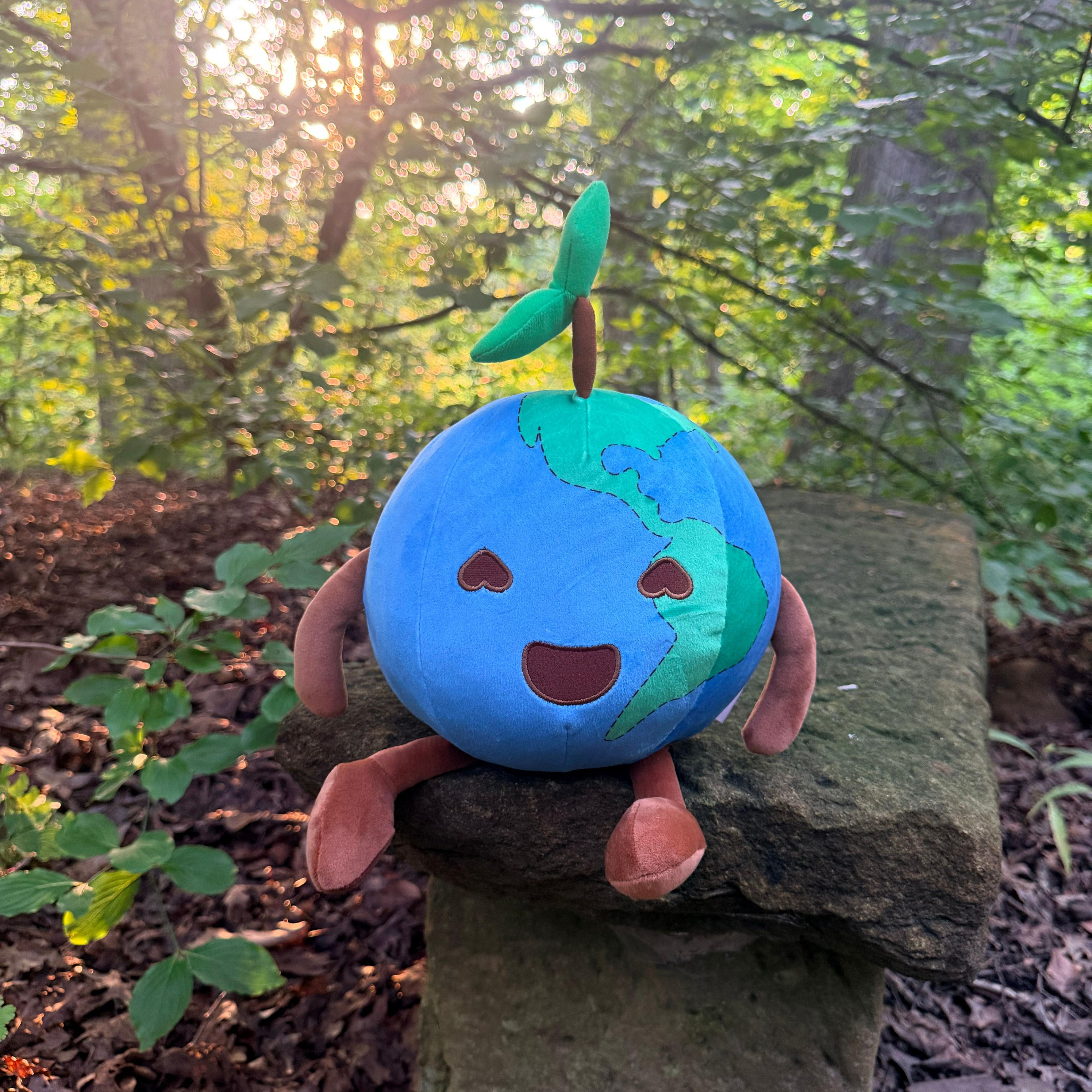 Smiling Earth plush toy with heart eyes and green sprout, sitting on a stone in a forest