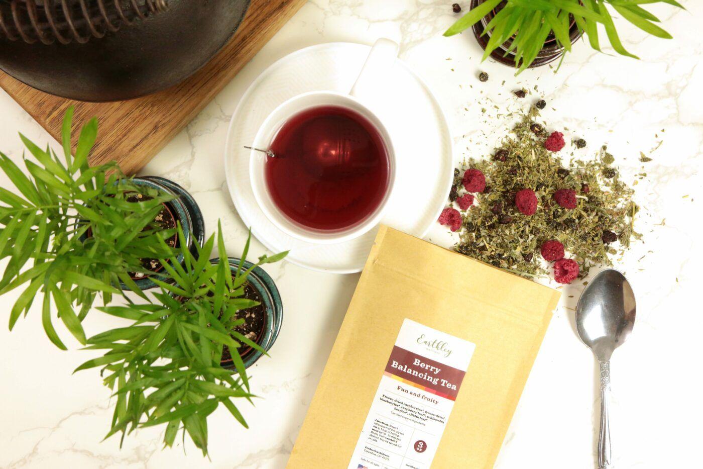 Earthley Berry Balancing Tea 3oz tan kraft resealable pouch with burgundy label on white marble with scattered green herb and red raspberry tea blend dark brown teapot white cup with dark red tea silver spoon and green plants in teal pots