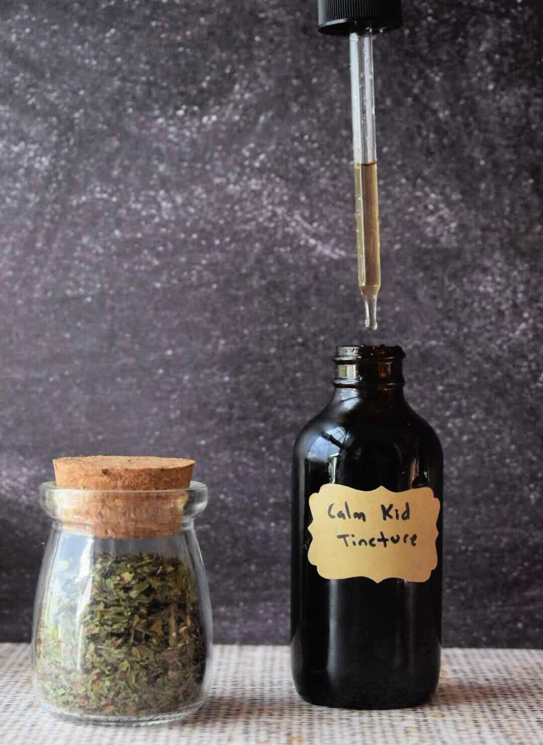 Glass dropper dispensing golden herbal tincture into dark bottle labeled "Calm Kid Tincture" next to jar of dried herbs