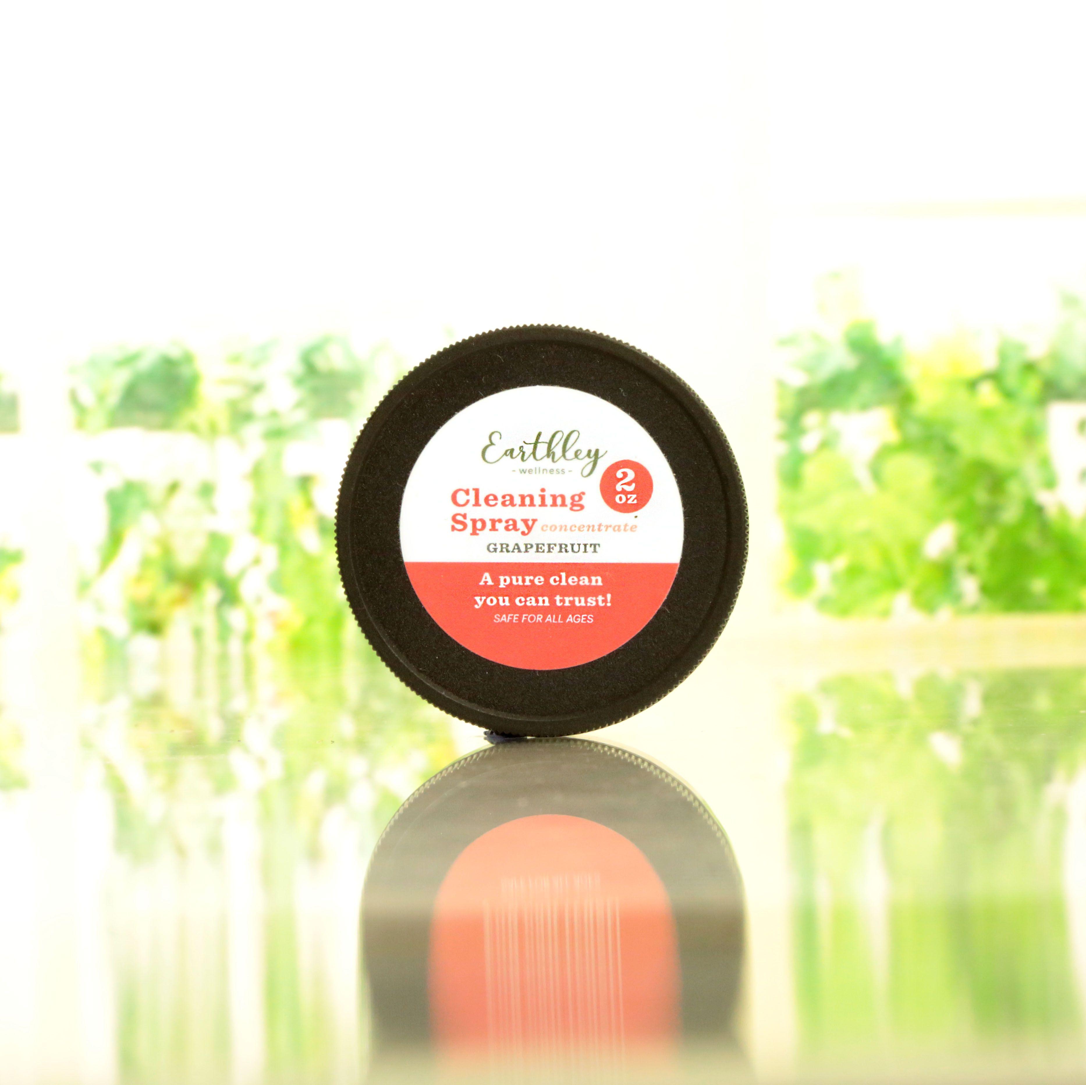 Earthley Cleaning Spray Concentrate Grapefruit 2oz black jar with coral and white label on reflective glass surface with green plants in background
