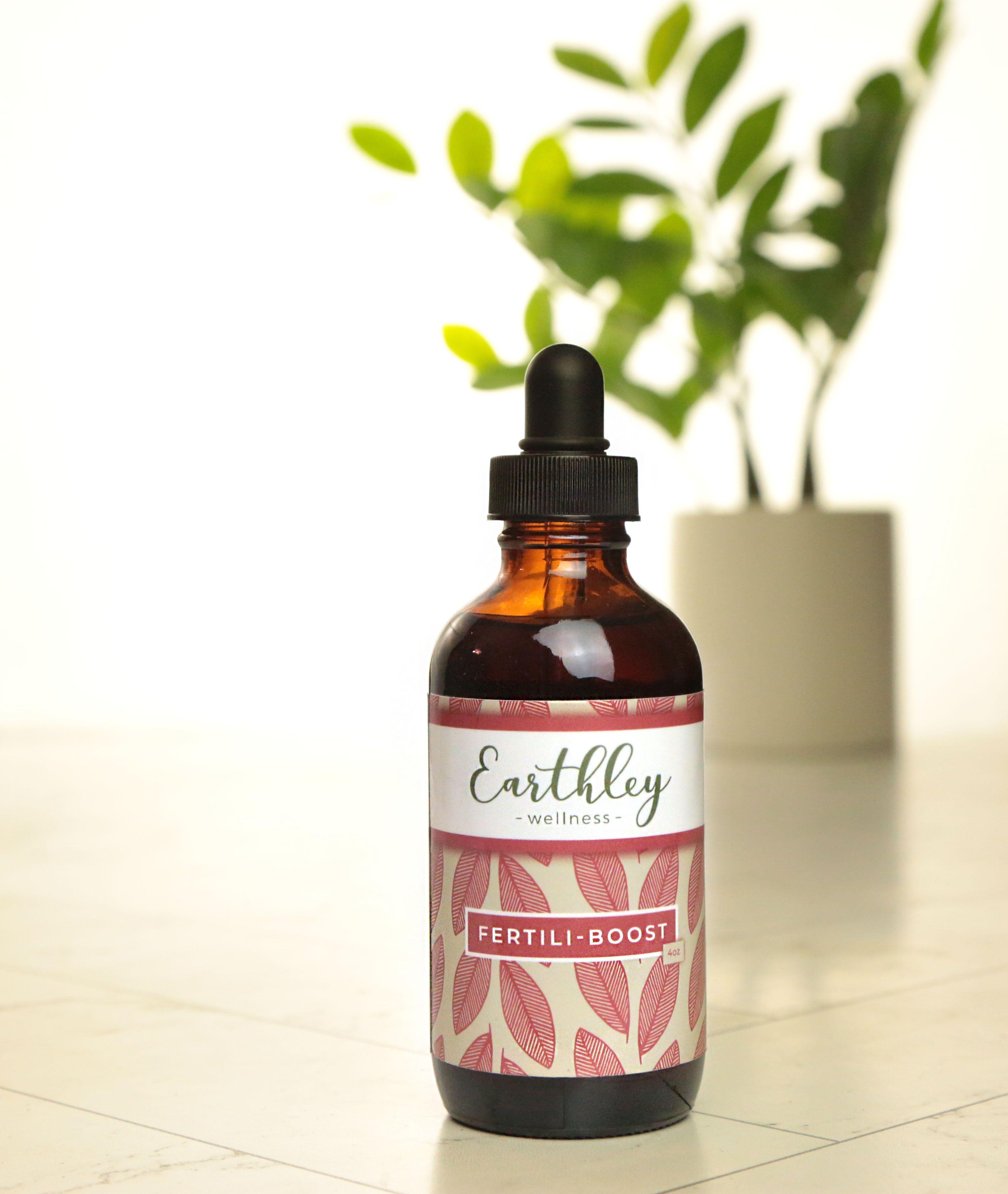 Fertili-Boost 4oz product photo showing amber glass dropper bottle with black cap and cream label with dusty rose mauve pink leaf pattern on white floor with green ZZ plant in cream white pot blurred in background