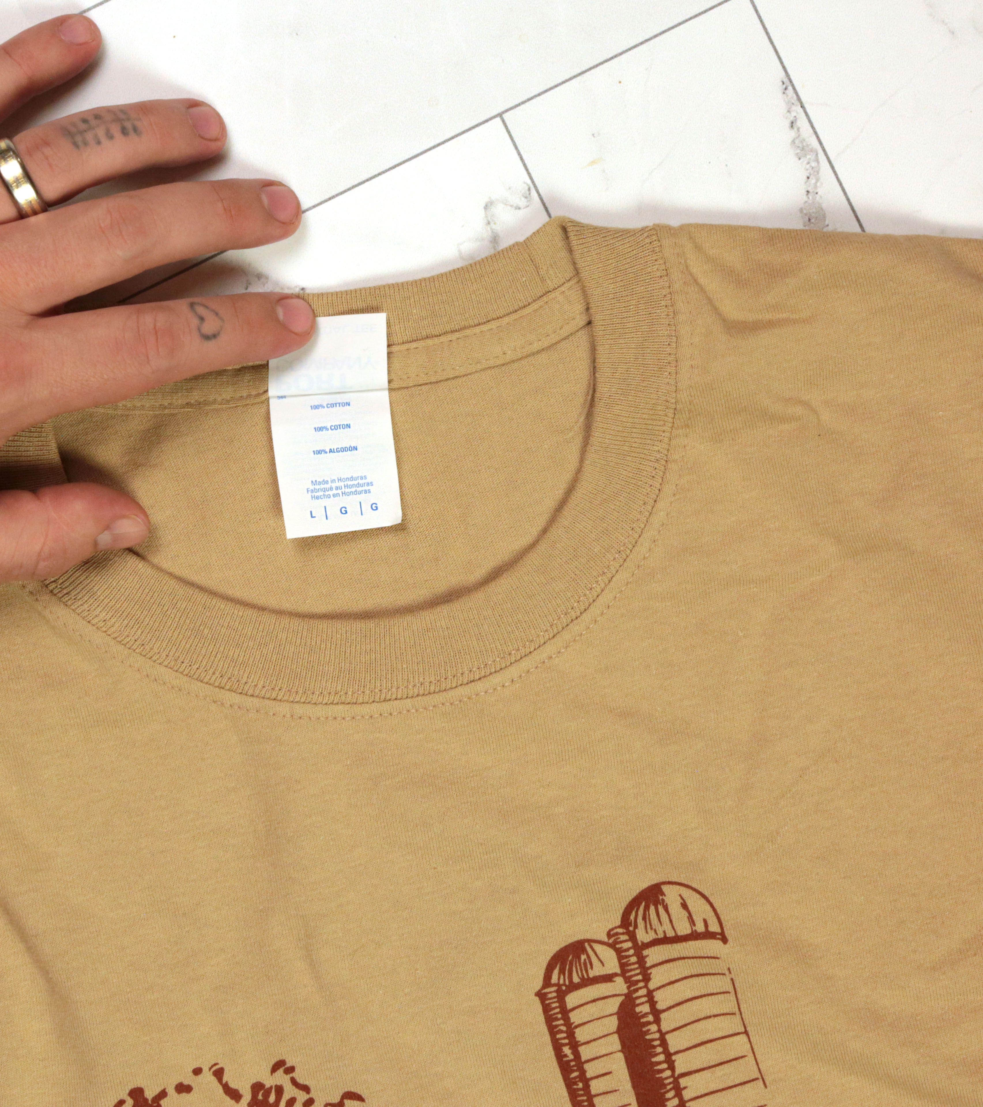 Close-up of tan gold Big Farms over Big Pharma t-shirt showing white care label and brown farm silo design with hand displaying gold ring