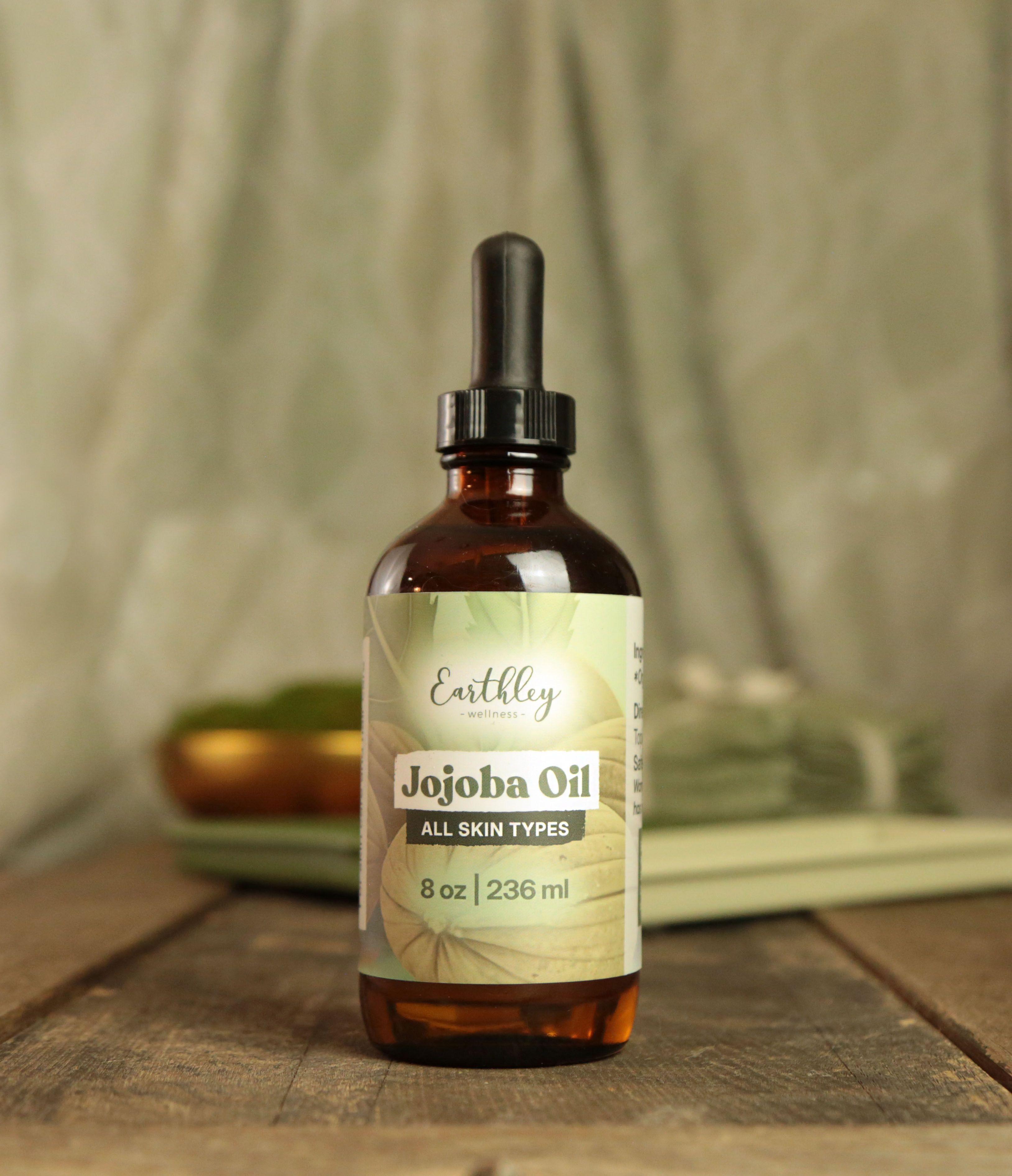 Earthley Jojoba Oil 8oz amber glass dropper bottle with sage green leaf pattern label for all skin types on rustic wood surface with spa accessories in background