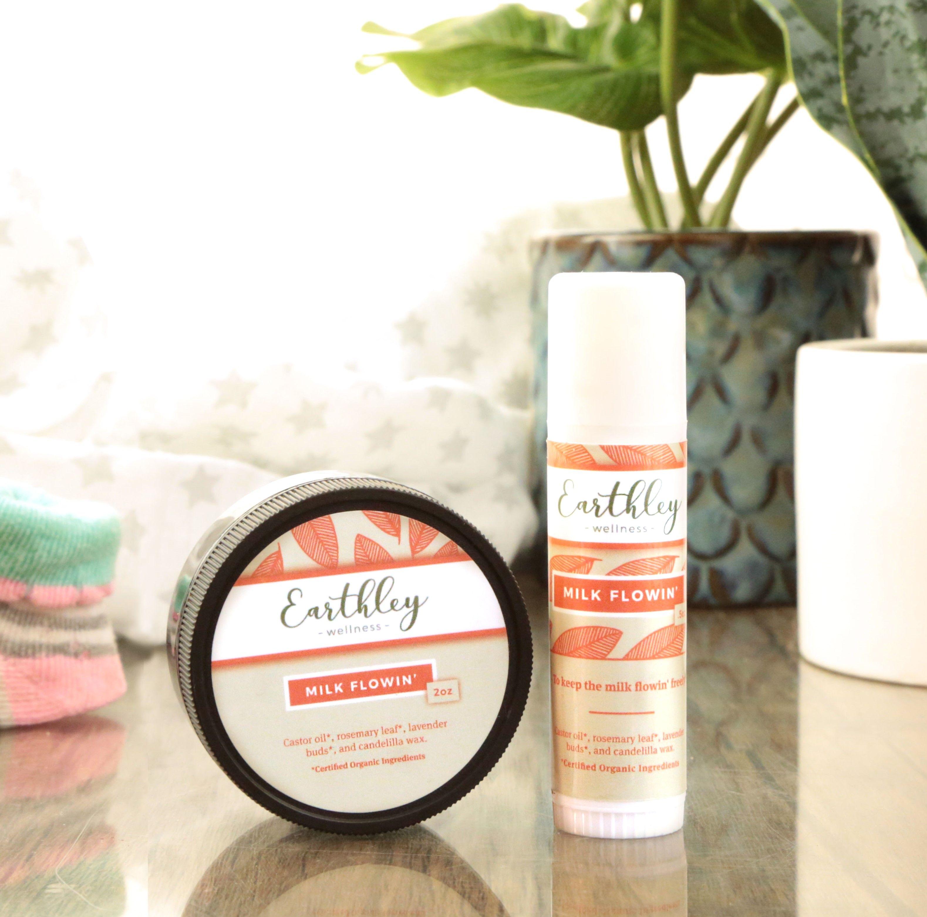 Earthley Milk Flowin breastfeeding support balm in 2oz black tin and twist-up stick with coral leaf pattern labels listing castor oil rosemary leaf lavender buds and candelilla wax with potted plant and baby socks in background
