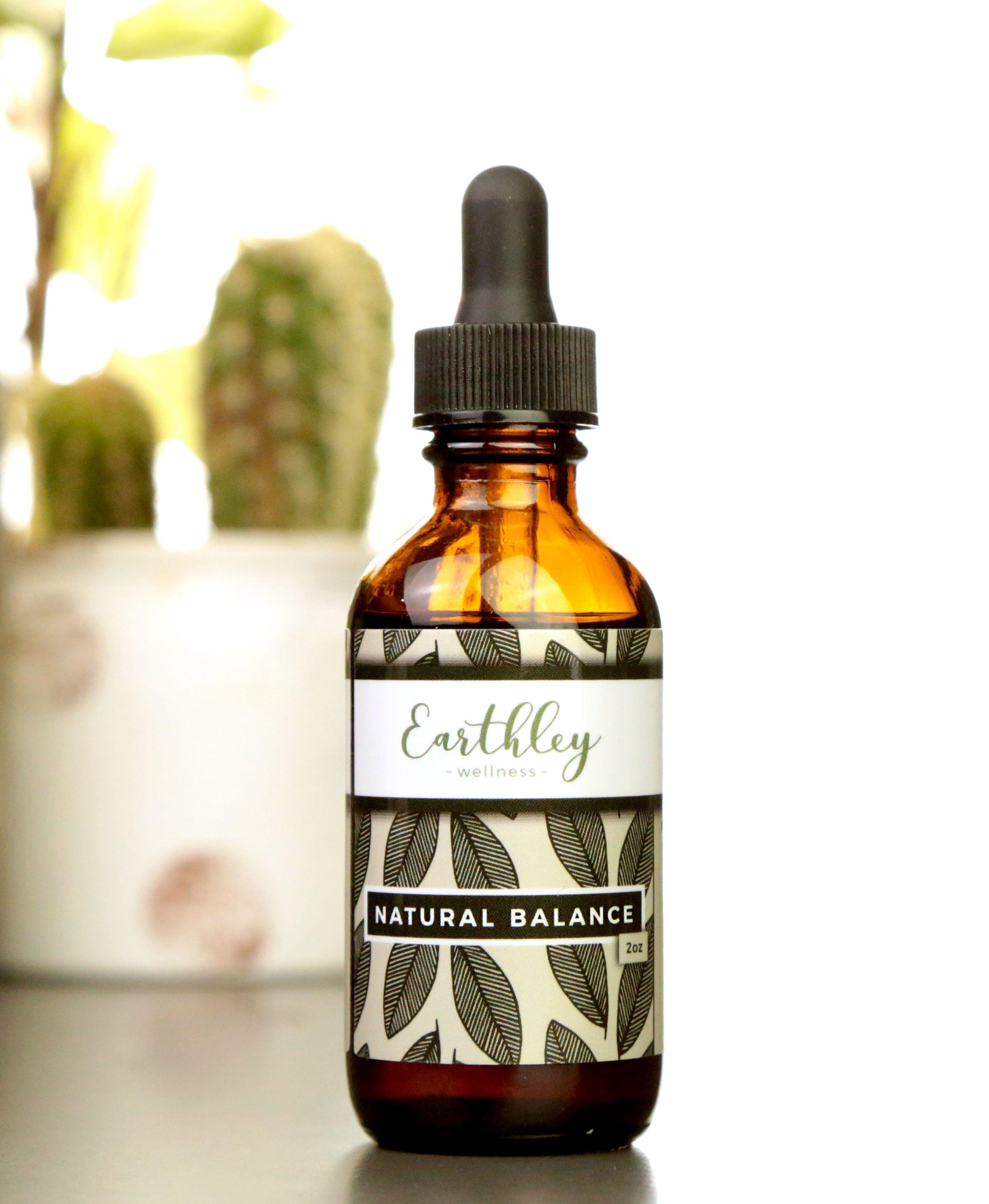 Natural Balance 2oz product photo showing amber glass dropper bottle with black cap and cream label with dark olive sage green leaf pattern on white surface with green cactus plant blurred in background