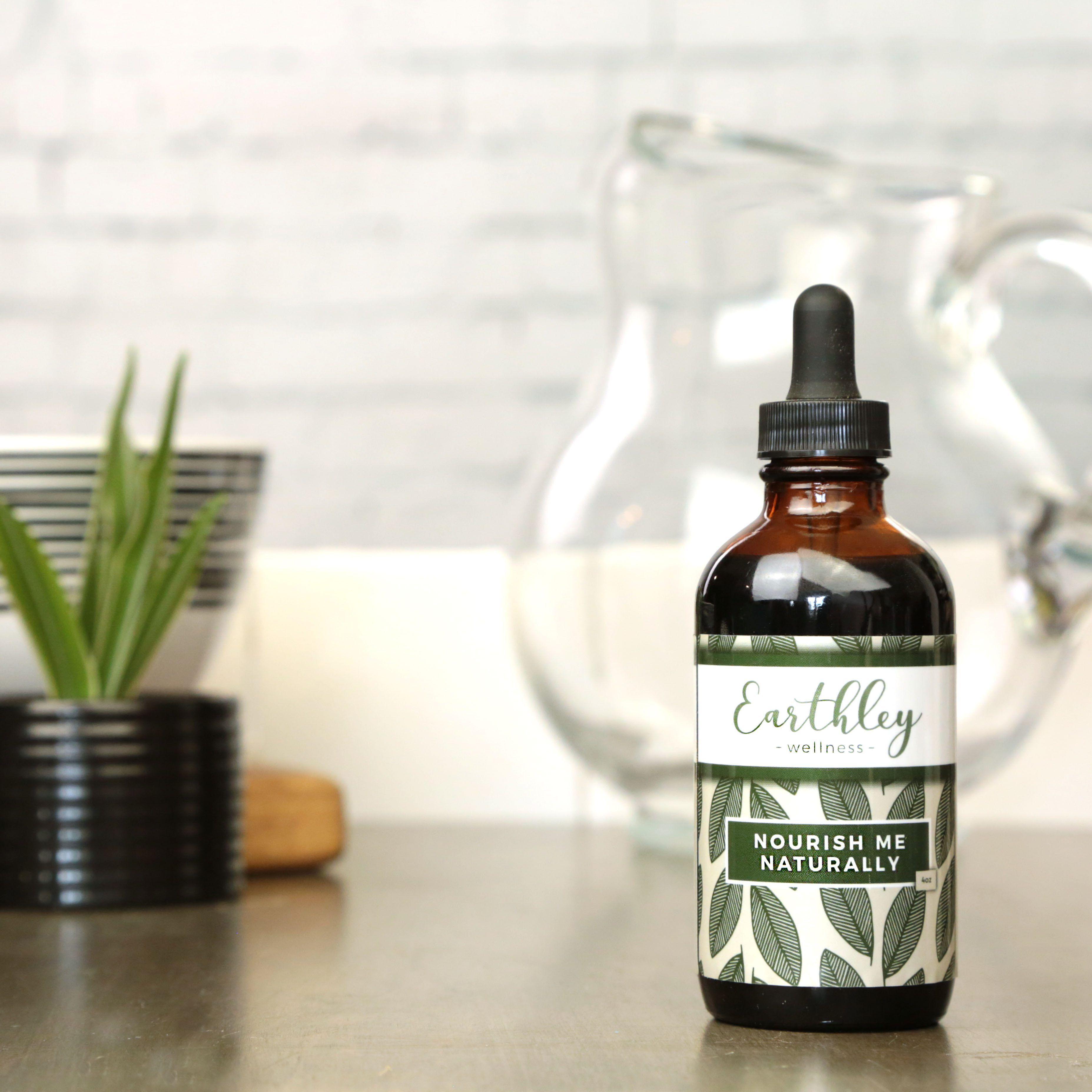 Earthley Nourish Naturally Herbal Multivitamin Extract amber glass dropper bottle with black cap and sage green leaf pattern label on white marble with green plants