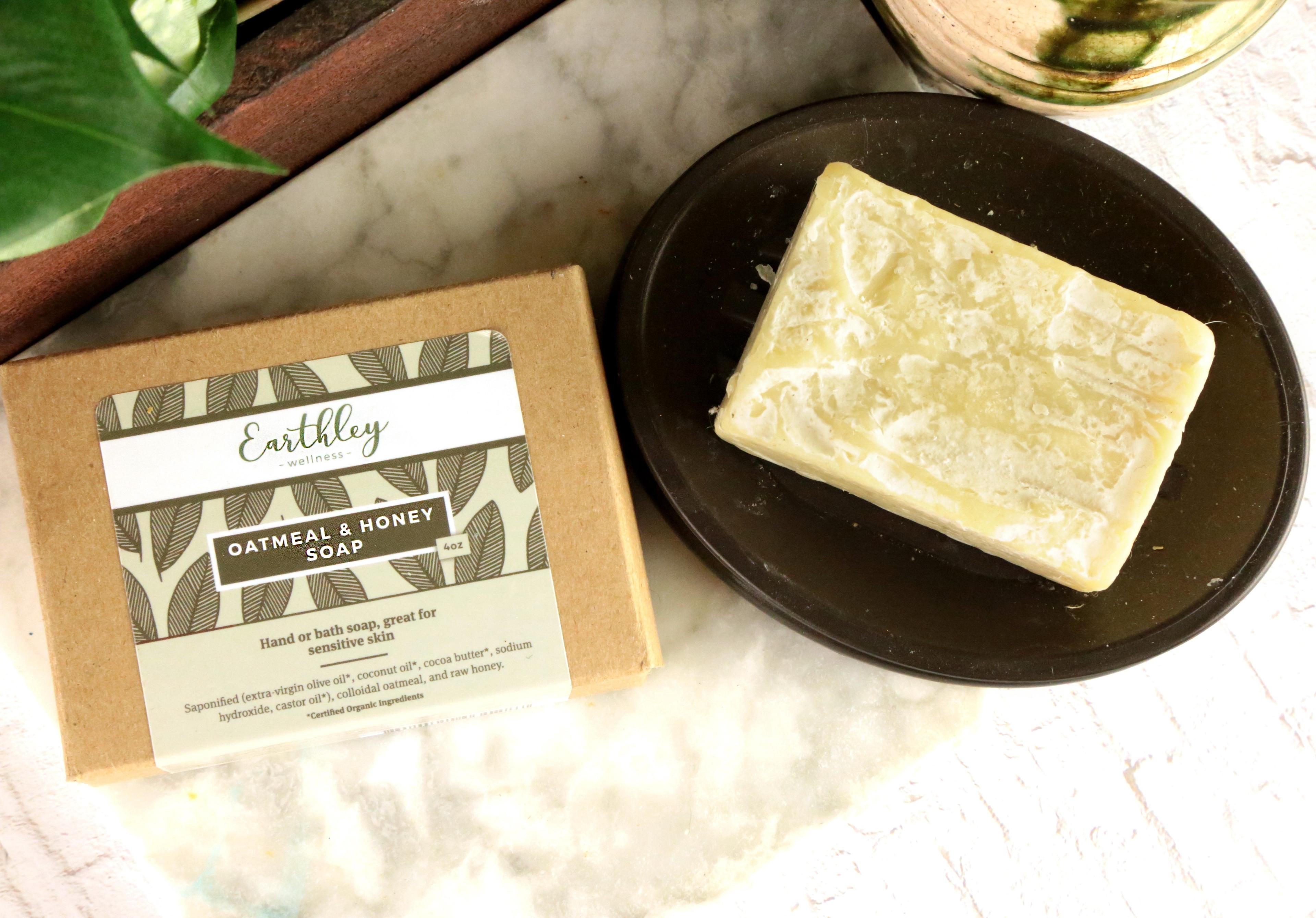Oatmeal and Honey Soap flatlay showing tan kraft box with cream label with olive gray leaf pattern and cream off-white soap bar on dark brown black ceramic dish on white marble surface with green leaves in dark wooden tray