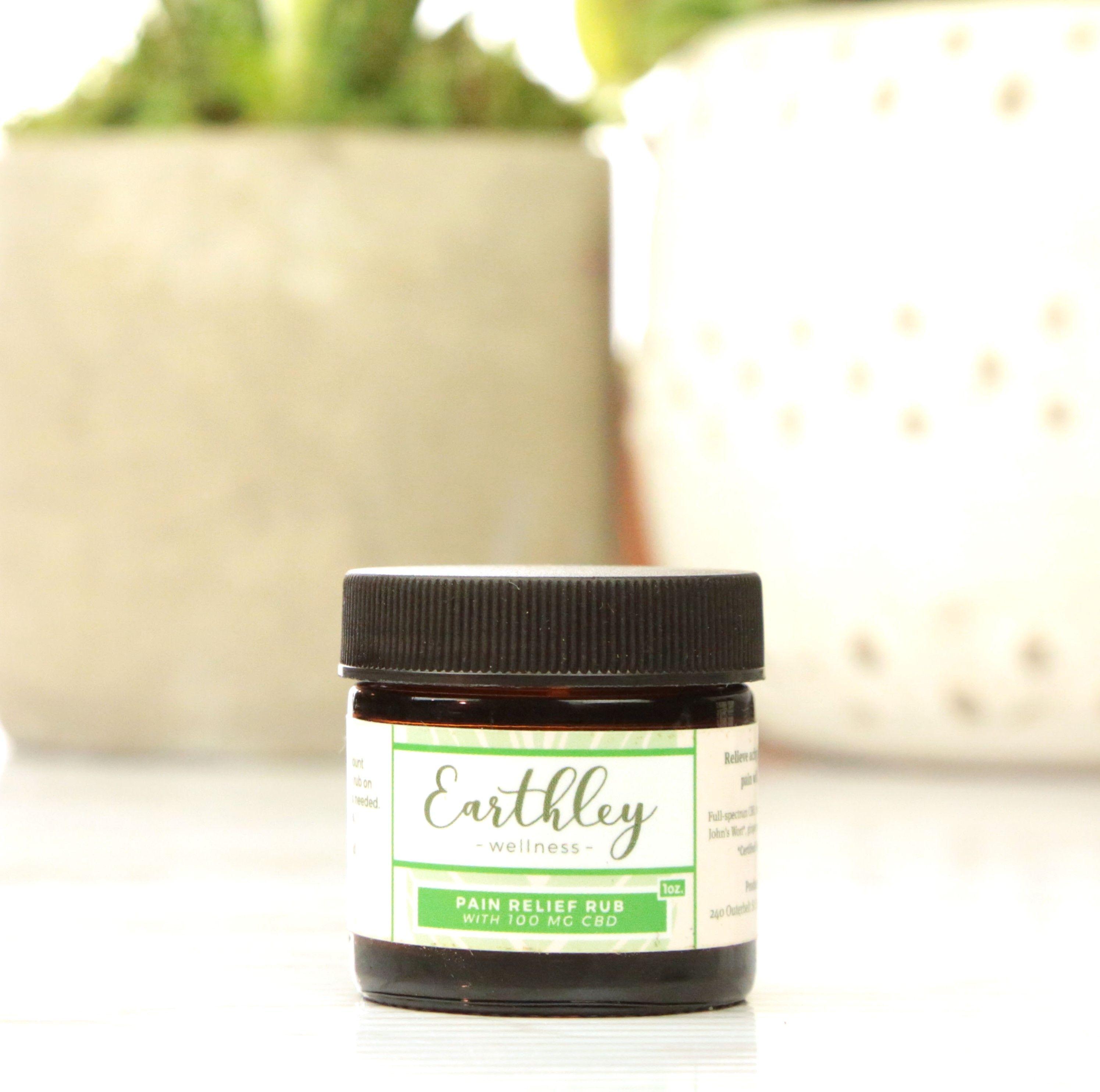 Earthley Pain Relief Rub 1oz amber glass jar with black lid and green stripe label showing CBD formula with 100mg CBD for topical pain relief with succulent plants in blurred background