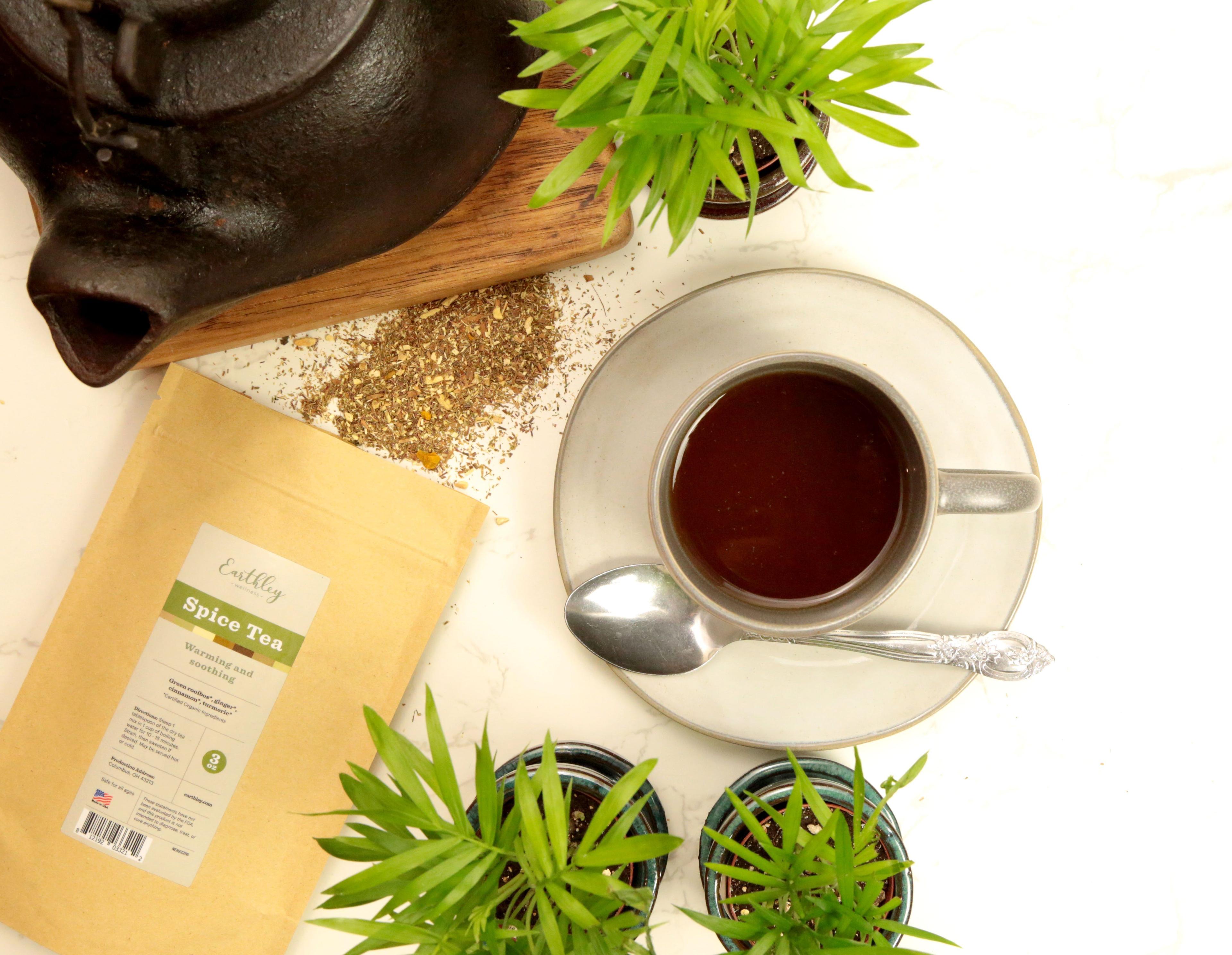 Earthley Spice Tea tan kraft resealable pouch with sage green label on white marble with dark brown cast iron teapot wooden cutting board scattered brown tea blend white teacup with silver spoon and green plants in teal pots