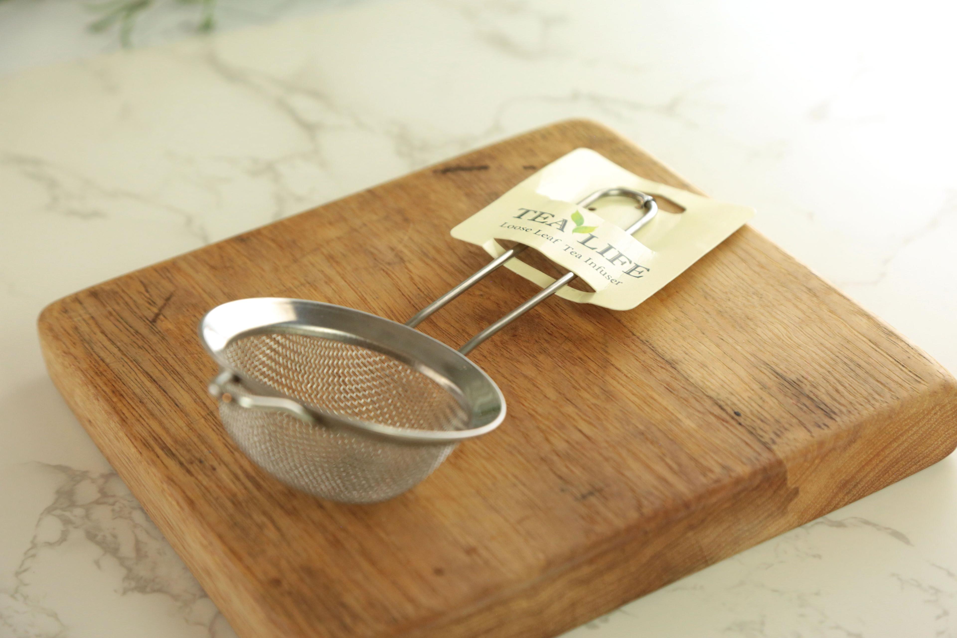 Tea Life stainless steel mesh tea infuser strainer with silver handle on wooden cutting board on white marble countertop