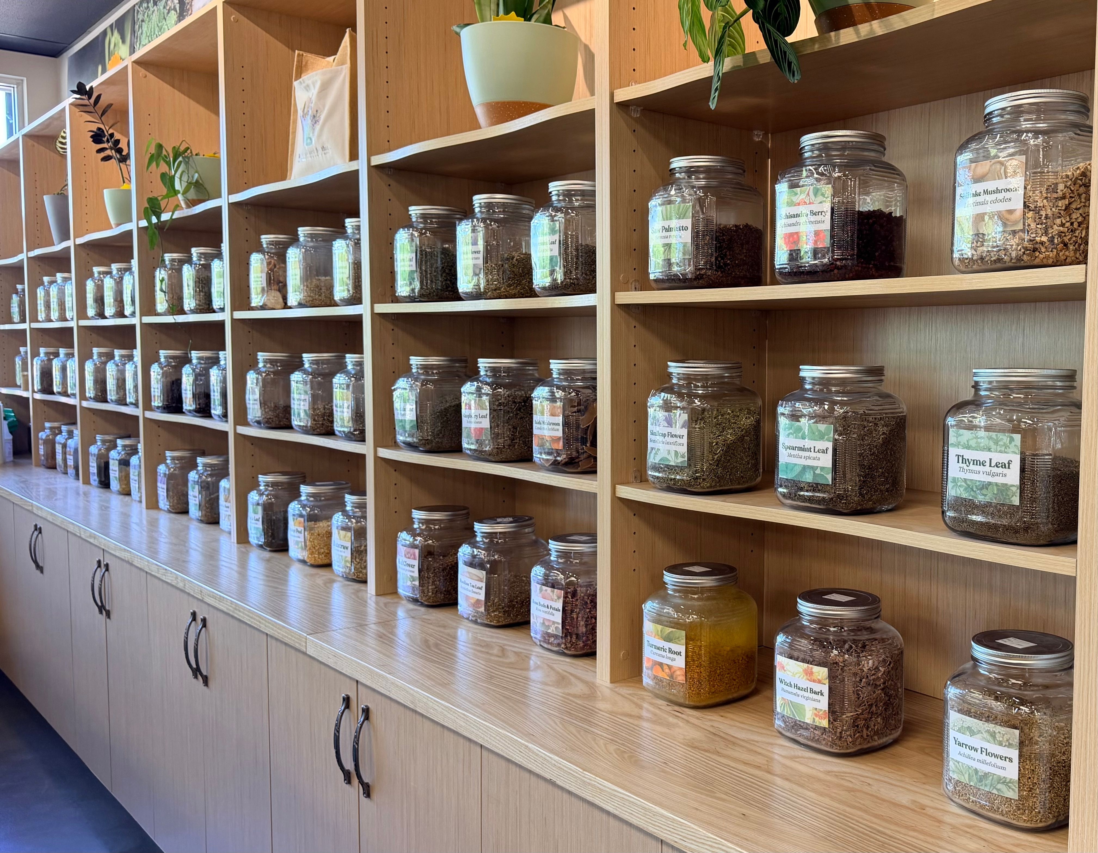 Wooden shelves lined with labeled glass jars of dried herbs including thyme, spearmint, and yarrow